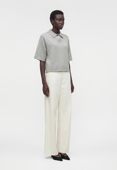 Γυναίκα που στέκεται, φοράει γκρι κορυφή με κοντά μανίκια και κολάρο, φαρδιά λευκά παντελόνια και μαύρα μυτερά παπούτσια, μπροστά σε απλό φωτεινό φόντο.