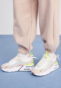 Fötter i beige och vita chunky sneakers med neongröna accenter och vita strumpor, stående på en blå matta med beiga sweatpants.