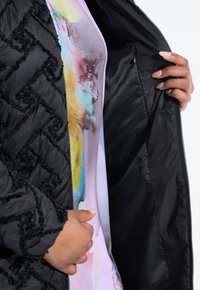 Svart vadderad jacka med texturerade detaljer, öppnad för att avslöja ett färgglatt, abstrakt mönstrat foder i pastellfärger.