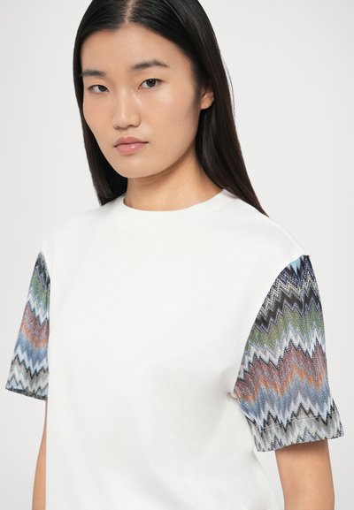 Λευκό μπλουζάκι με πλεκτά μανίκια σε μπλε, πράσινο, πορτοκαλί και μαύρα ζιγκ-ζαγκ σχέδια. Λαιμόκοψη crew και κοντά μανίκια. Λεπτή υφή.