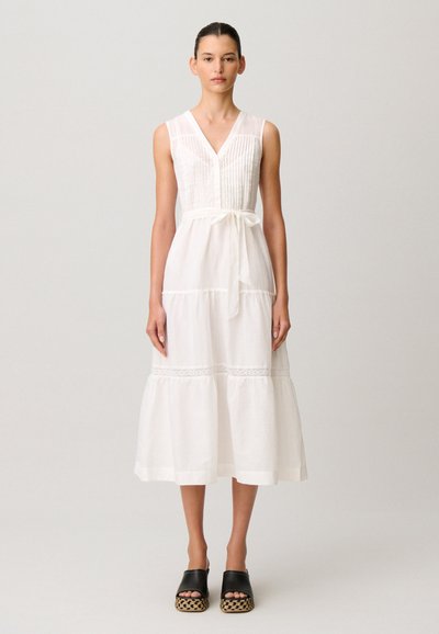 Donna in piedi che indossa un vestito midi bianco senza maniche con scollo a V, cintura in vita e gonna a balze, abbinato a sandali neri con plateau.