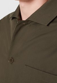 Camicia verde oliva con colletto, chiusura a un bottone e taschino sul petto. Il tessuto ha una finitura liscia e strutturata.