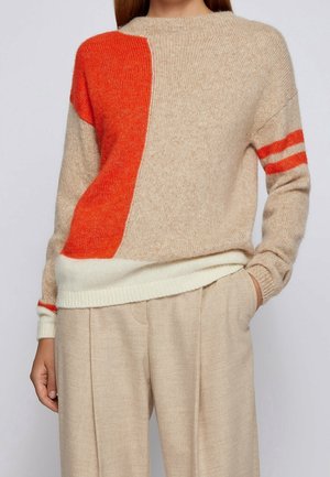 Woman wearing a beige knit sweater with an orange block on the left side and orange stripes on the right sleeve, paired with beige trousers.