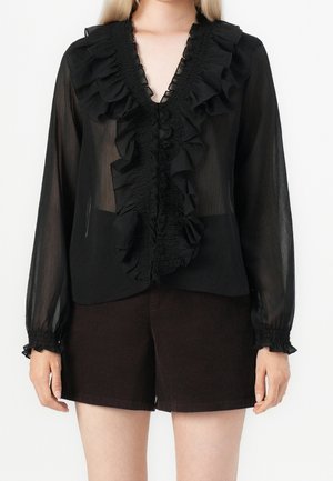 Kobieta ubrana w czarną przezroczystą bluzkę z falbaniastym dekoltem w kształcie litery V i mankietami, w połączeniu z ciemnobrązowymi szortami, stojąca na jednolitym tle.