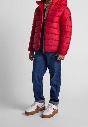 Doudoune rouge avec capuche, matelassage horizontal et écusson. Portée sur une chemise rayée, associée à un jean bleu et des baskets blanches.