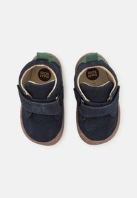 Sapatos de camurça azul-marinho com detalhes em verde e tiras de Velcro. Ponta arredondada, sola de borracha castanha. O interior apresenta o logótipo da marca e um forro suave.