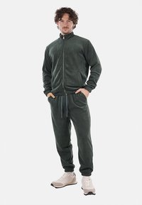 Tuta in pile verde scuro con giacca con zip e pantaloni con vita elastica, caratterizzato da una texture morbida e polsini a costine. Indossato con scarpe da ginnastica leggere.