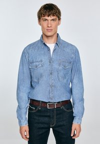 Camisa de denim azul claro de mangas largas, con botones a presión y dos bolsillos en el pecho, llevada sobre una camiseta blanca, combinada con jeans oscuros y un cinturón marrón.