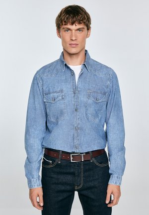 Camisa de denim azul claro de mangas largas, con botones a presión y dos bolsillos en el pecho, llevada sobre una camiseta blanca, combinada con jeans oscuros y un cinturón marrón.