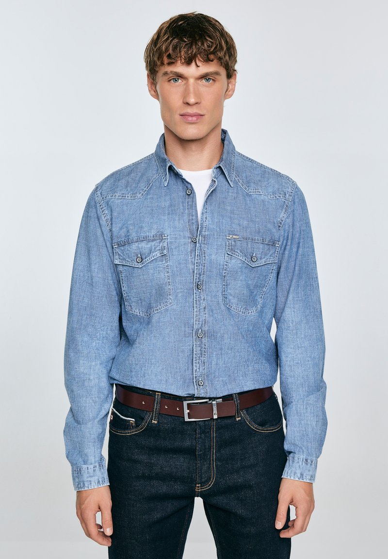 Camisa de denim azul claro de mangas largas, con botones a presión y dos bolsillos en el pecho, llevada sobre una camiseta blanca, combinada con jeans oscuros y un cinturón marrón.