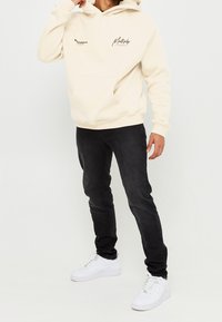 Beige hoodie met voorzak en gedrukt tekst, gecombineerd met zwarte skinny jeans en witte sneakers. Eenvoudig, casual ontwerp.
