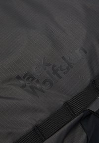 Černá nylonová tkanina s texturovaným vzorem, obsahující vyvýšené logo "Jack Wolfskin" v tmavé barvě a detail s černým popruhem z tkaniny.