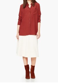 Camisa roja de botones con dos bolsillos en el pecho, combinada con una falda blanca. Botines de ante marrón completan el conjunto.