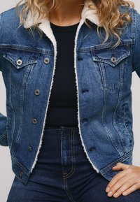 Džínsová bunda s lehkým límcem z umělé kožešiny, dvěma předními kapsami a knoflíkovým zapínáním. Modrý odstín s jemným vyblednutím a detaily v prošívání.