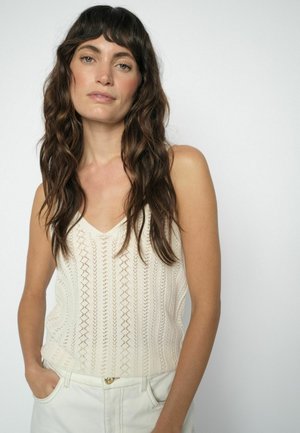 Donna con capelli castani lunghi e ondulati che indossa un top bianco senza maniche in maglia e pantaloni bianchi, in piedi con le mani nelle tasche contro uno sfondo semplice.