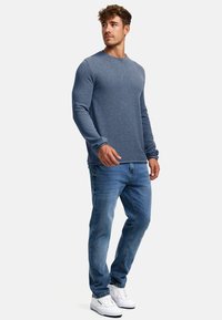 Sininen pitkähihaisen paita, jossa on pyöreä kaula-aukko, yhdistettynä vaaleansinisiin farkkuihin ja valkoisiin lenkkitossuihin. Kangas näyttää pehmeältä ja tekstuuriltaan erikoiselta.