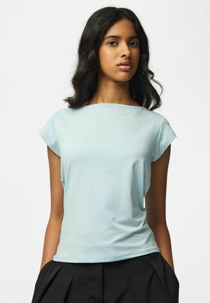 Jeune femme aux cheveux foncés portant un haut bleu clair à manches courtes et un pantalon noir, debout devant un fond blanc uni.