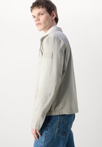 Lys beige jakke, lett materiale, avslappet passform, med ribbestrikket krage, elastiske ermer og et logo-patch på venstre erme.