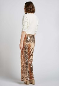 Pantalons wide-leg en sequins rose doré avec une texture lisse, associés à un pull crème, présentés de dos. Des chaussures de couleur neutre complètent le look.