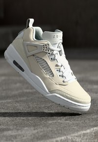 Beige színű sportcipők texturált bőr és hálós panelekkel, fehér középtalppal és fűzőkkel, valamint látható légpárnázással a talpon.