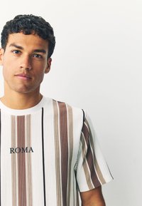 White ribbed t-shirt featuring vertical brown and beige stripes. "ROMA" embroidered in black at the chest. Short sleeves and crew neck design.