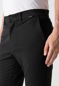 Schwarze Hosen mit schmalem Schnitt, die eine glatte Textur, einen flachen Bund und seitliche Taschen aufweisen. Knopfverschluss mit Calvin Klein-Logo am Bund.