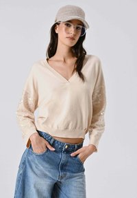 Maglione beige con scollo a V e maniche lunghe con dettagli in paillettes, abbinato a jeans di jeans a vita alta. Cappello e occhiali non inclusi.