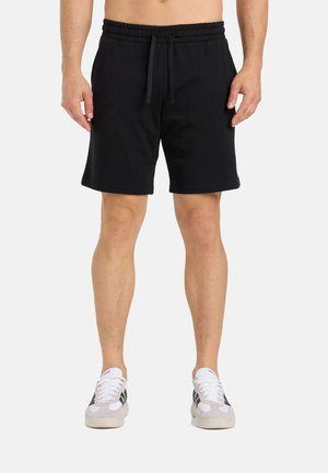 Uomo che indossa pantaloncini neri con coulisse e scarpe da ginnastica bianche con strisce nere, in piedi davanti a uno sfondo semplice.