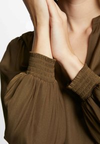 Blusa marrone con polsini arricciati e maniche voluminose, caratterizzata da una texture morbida e fluida che esalta la silhouette del capo.