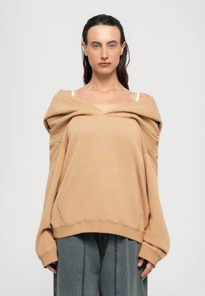 Kvinna med mörkt hår bär en oversize beige av-axel sweatshirt över vita remmar och lösa grå byxor, stående mot en enkel ljus bakgrund.