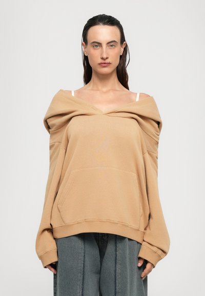 Vrouw met donker haar draagt een oversized beige off-shoulder sweatshirt over witte bandjes en losse grijze broek, staand tegen een effen lichte achtergrond.