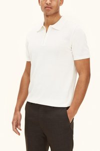 Valkoinen, lyhythihainen poolopaita, jossa on kaulus ja puolipitkä napitus, teksturoitua kangasta, yhdistettynä tummiin räätälöityihin housuihin.