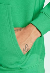 Sudadera verde con una tela texturizada. Cuenta con un bolsillo tipo canguro y mangas con puños. Tatuaje visible en la mano que descansa sobre el bolsillo.