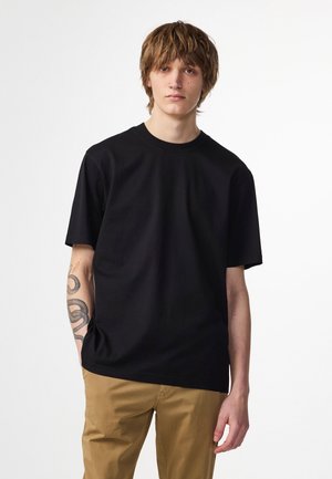 Schwarzes Baumwoll-T-Shirt mit kurzen Ärmeln, Rundhalsausschnitt und lockerer Passform. Getragen mit khakifarbenen Hosen, mit einem Tattoo am Arm.