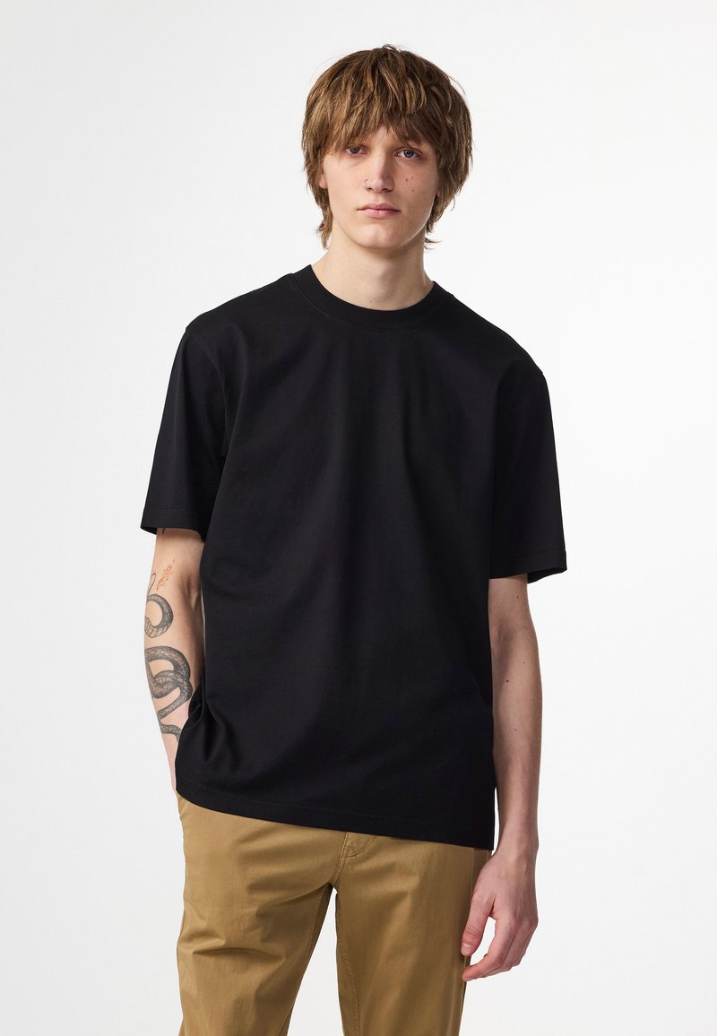 Svart bomullströja med korta ärmar, rund hals och en avslappnad passform. Bärs med beige byxor och har en tatuering på armen.