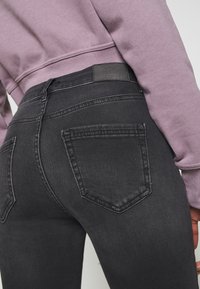 Jeans de mezclilla negra con un diseño ajustado, que cuentan con dos bolsillos traseros y un parche de cuero en la cinturilla. Acompañados de un sudadera de color púrpura claro.