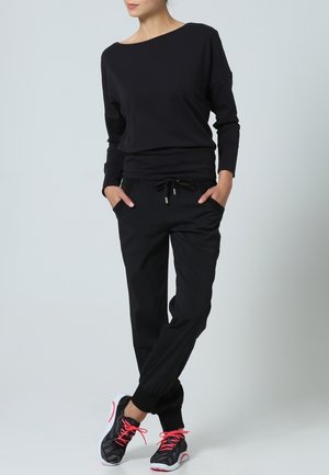 Žena stojící s rukama v kapsách, oblečená v černém dlouhém topu, černých teplácích a černých sportovních botách s růžovými tkaničkami.