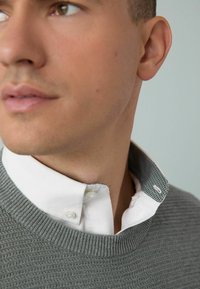 Bărbat cu ten deschis, purtând un pulover gri din tricot peste o cămașă albă cu guler colorat, uitându-se pe partea laterală, pe un fundal neutru.