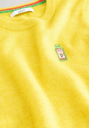 Dzeltena džemperis ar zaļu un sarkanu svītru apkakli un izšūtu karstā mērces pudeles plāksteri uz krūtīm, ar uzrakstu "KARSTĀ LIETA".