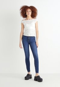 Vit, strukturerad topp med avtagna axlar och rynkdetalj; ihop med mörkblå, figurnära jeans och svarta klackskor.