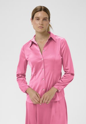 Kobieta o jasnobrązowych włosach związanych z tyłu, ubrana w długorękawową, różową satynową bluzkę, stojąca z rękami złożonymi przed sobą na jednolitym tle.