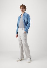 Camisa de mezclilla azul clara sobre una camiseta sin mangas gris, combinada con pantalones cargo color blanco roto con bolsillos y zapatillas blancas con detalles grises.