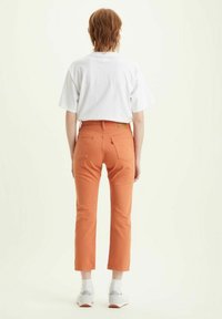 Jeans arancioni a vita alta, con tasche posteriori, passanti per la cintura e dettagli di cucitura sottili. Abbinati a una t-shirt bianca a maniche corte.