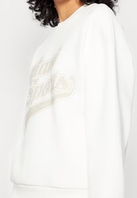 Vit sweatshirt med en avslappnad passform, med en texturerad grafisk tryck i beige. Materialet verkar mjukt med ribbade muddar och nederkant.