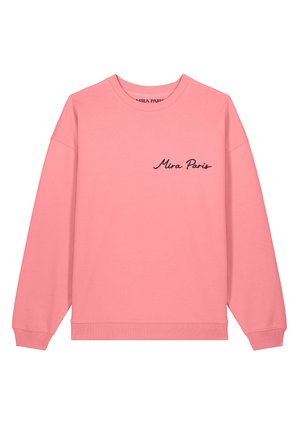 Femme blonde face à gauche portant un sweat rose ample avec le texte « Nin Paris » et un jean bleu, debout devant un fond clair uni.