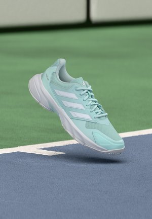 Baskets Adidas bleu clair avec des bandes blanches lévitant au-dessus de la surface bleue et verte d'un court de tennis.