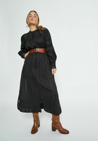 Black, long-sleeved dress with subtle textures and lace accents, worn with a brown leather belt. Knee-high brown boots complete the look.