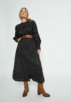 Abito nero a maniche lunghe con trame sottili e dettagli in pizzo, indossato con una cintura in pelle marrone. Stivali marroni fino al ginocchio completano il look.