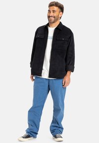 Giacca di velluto a coste nera con due tasche sul petto, indossata sopra una t-shirt bianca. Abbinata con pantaloni blu e sneakers nere. Vestibilità casual.