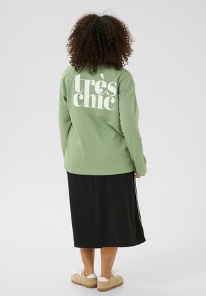 Green long-sleeve sweatshirt featuring large white text "très chic" on the back, paired with a black knee-length skirt and beige footwear.